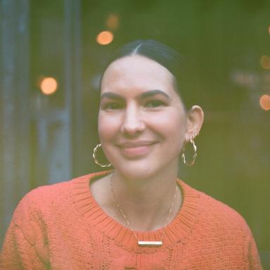 Linda smiles at camera with her black hair tied up and wearing gold hoop earrings and a orange jumper